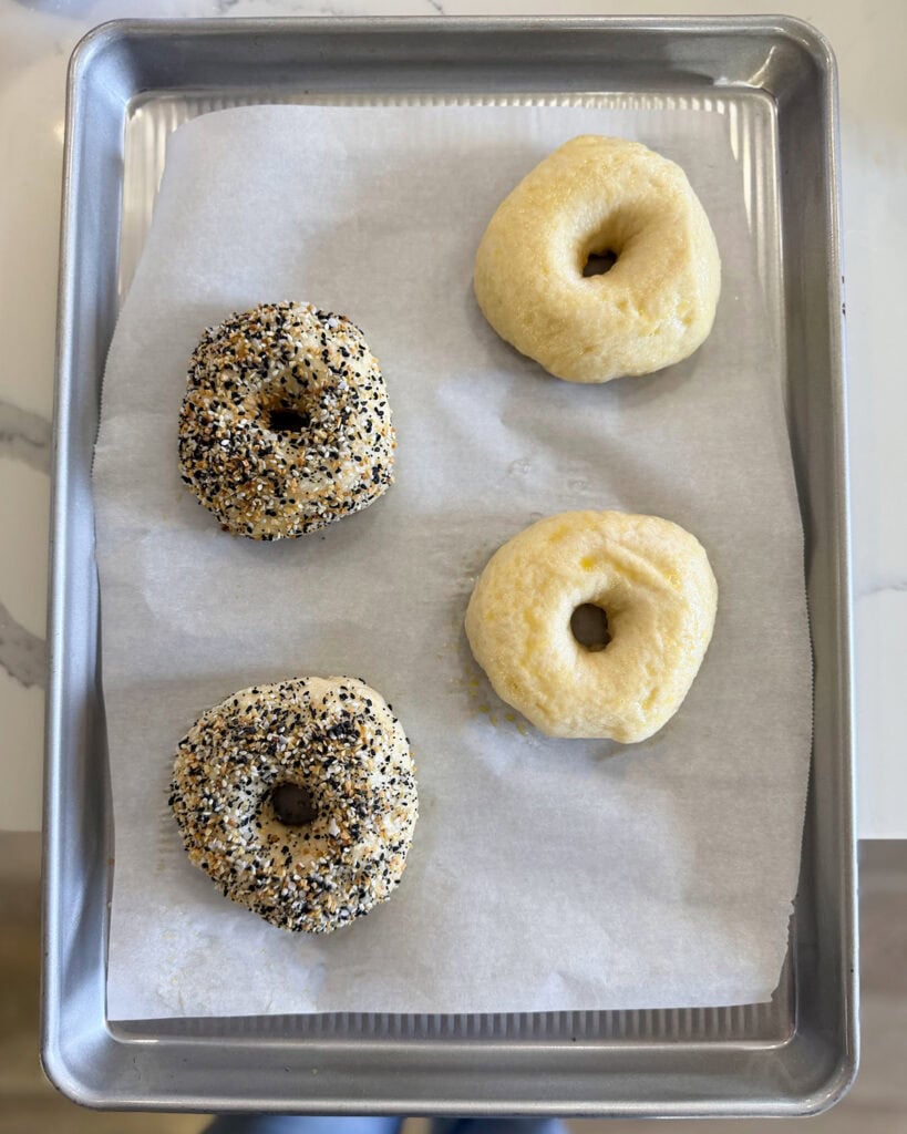 Four bagels on a sheet pan before baking, two are covered in everything bagel seasoning.