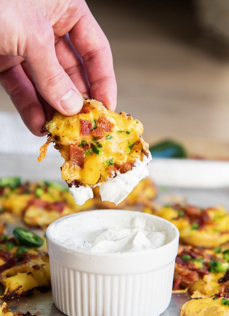 A hand dipping a crispy smashed potato topped with cheese and bacon in a bowl of sour cream.