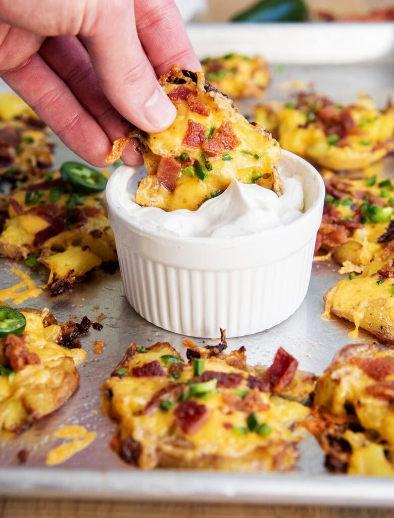A hand dipping a crispy smashed potato topped with cheese and bacon in a bowl of ranch dip.