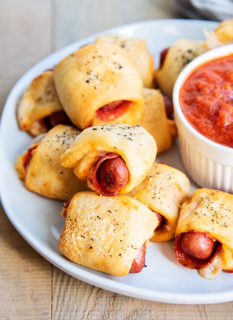 A pile of Pizza pigs in a blanket on a plate next to a bowl of pizza sauce.