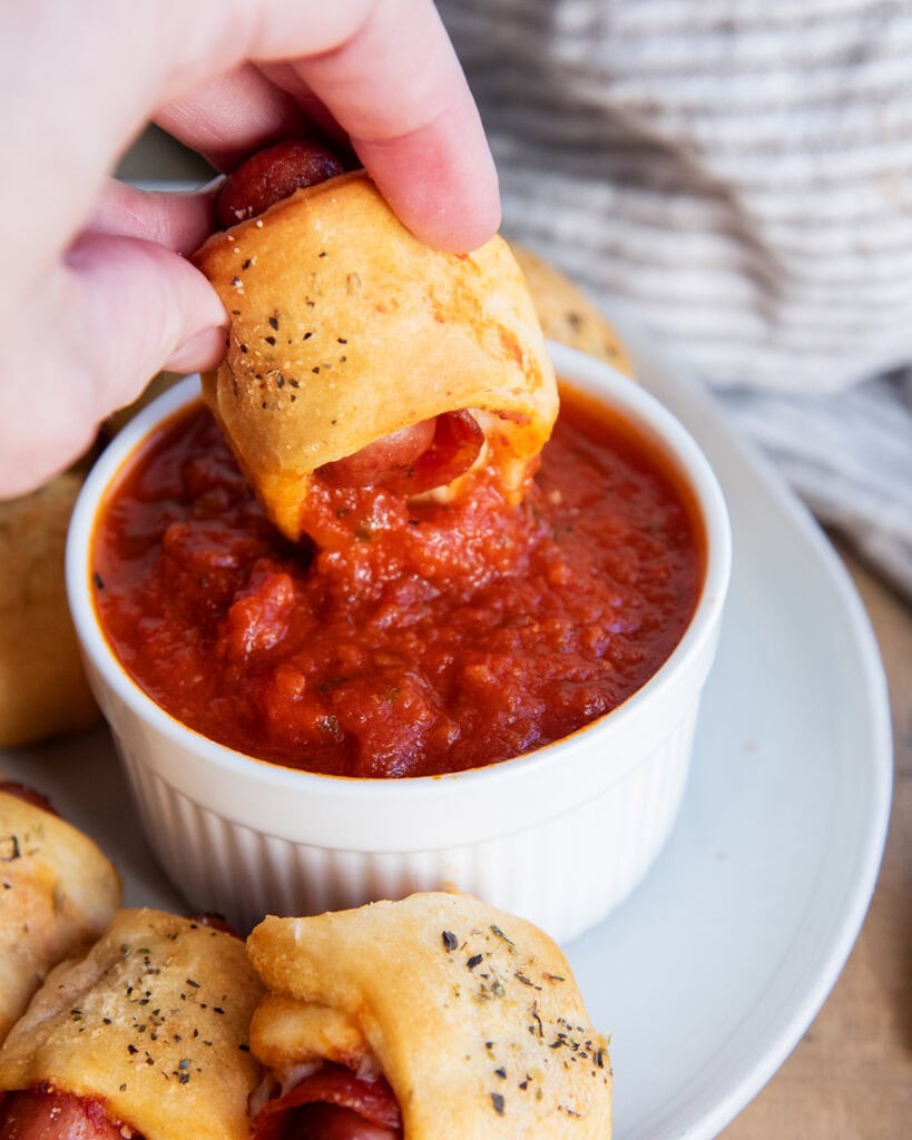 A hand dipping a pizza pigs in a blanket into pizza sauce.