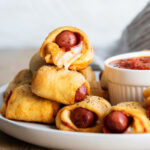 A pile of Pizza pigs in a blanket on a plate next to a bowl of pizza sauce.