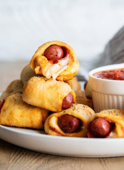 A pile of Pizza pigs in a blanket on a plate next to a bowl of pizza sauce.