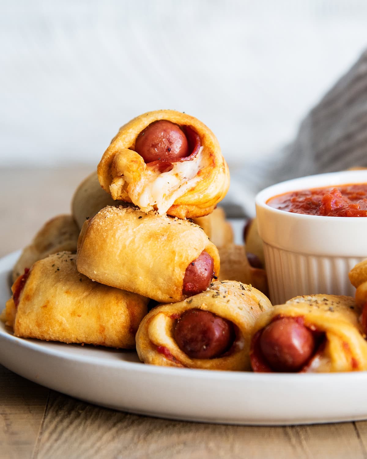 A pile of Pizza pigs in a blanket on a plate next to a bowl of pizza sauce.
