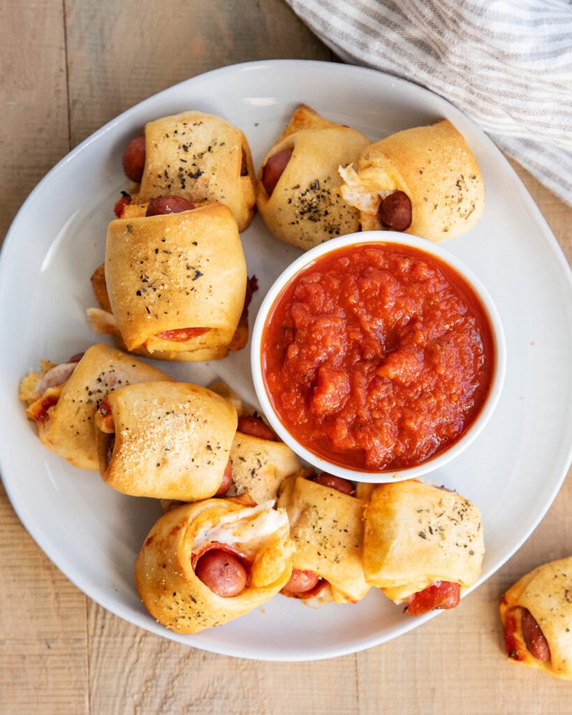 A pile of Pizza pigs in a blanket on a plate next to a bowl of pizza sauce.