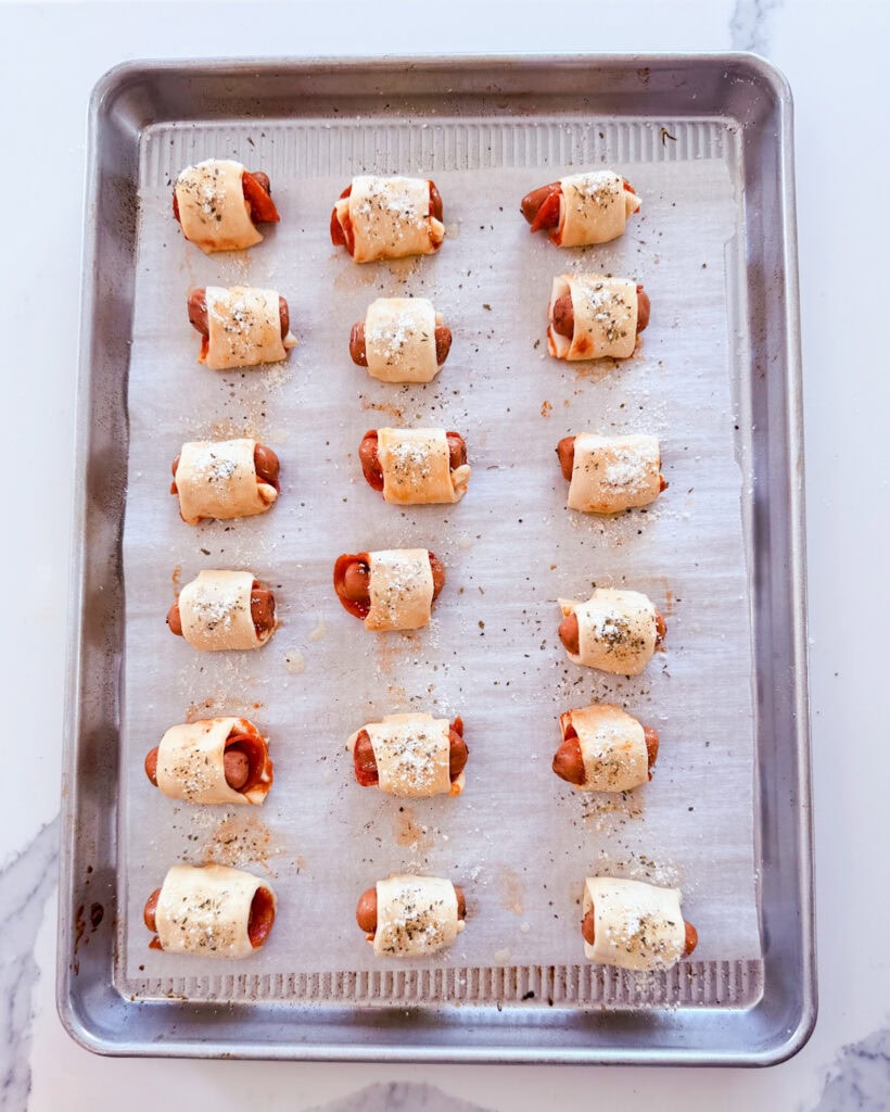 Unbaked pigs in a blanket on a cookie sheet, topped with Italian seasoning and parmesan.