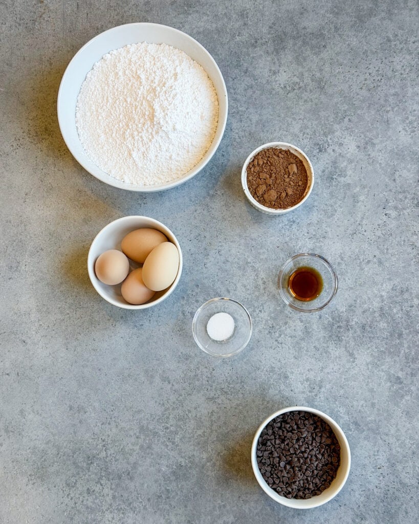 The ingredients needed to make flourless chocolate cookies, including powdered sugar, egg whites, cocoa powder, vanilla, salt, and chocolate chips. 