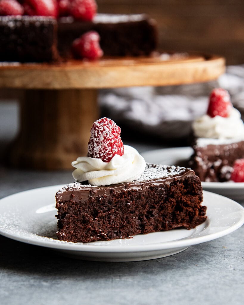 A slice of flourless chocolate cake topped with chocolate ganache, whipped cream, and a raspberry on a plate.