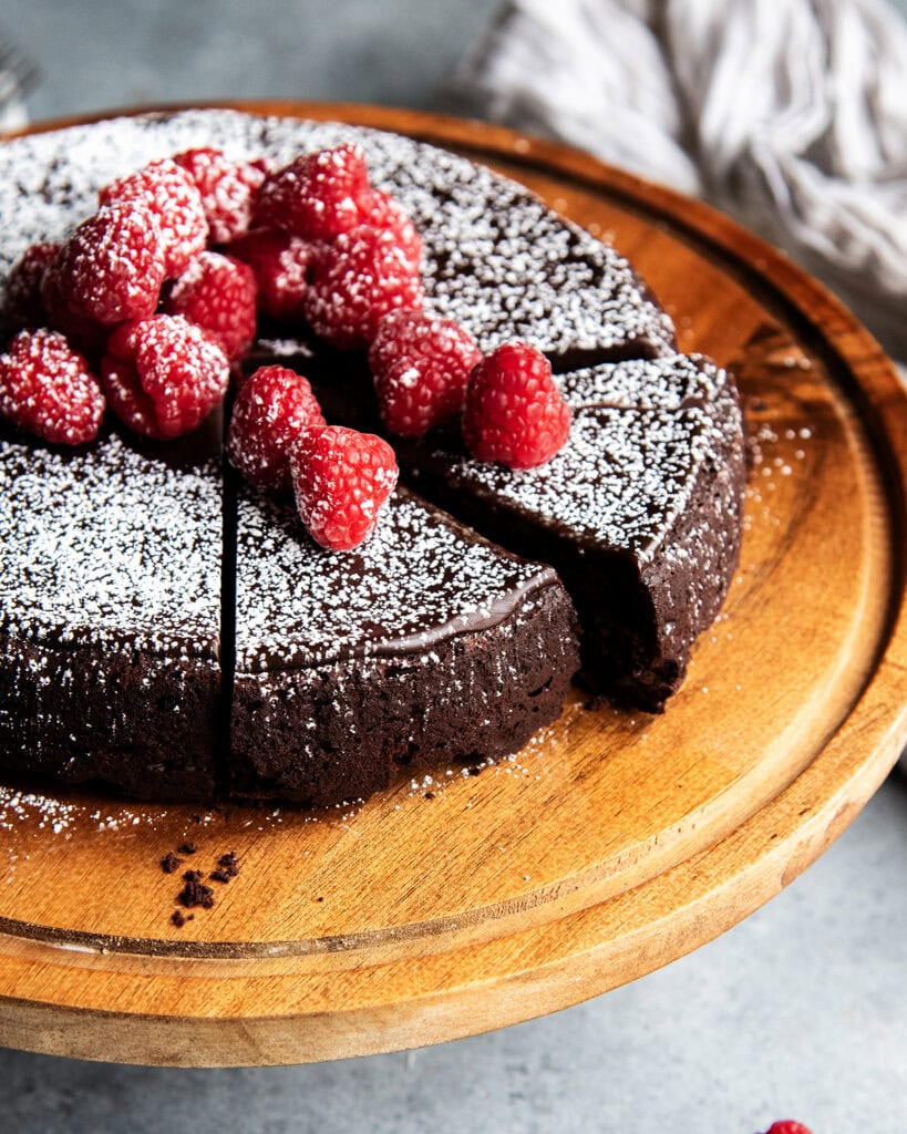 Flourless chocolate cake topped with chocolate ganache on a wooden cake stand.