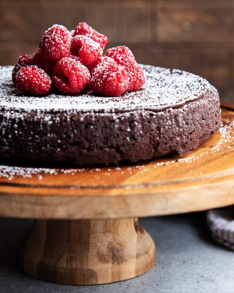 Flourless chocolate cake topped with chocolate ganache on a wooden cake stand.