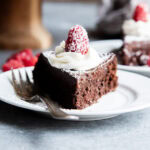 A slice of flourless chocolate cake topped with chocolate ganache, whipped cream, and a raspberry on a plate.