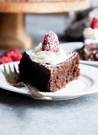 A slice of flourless chocolate cake topped with chocolate ganache, whipped cream, and a raspberry on a plate.