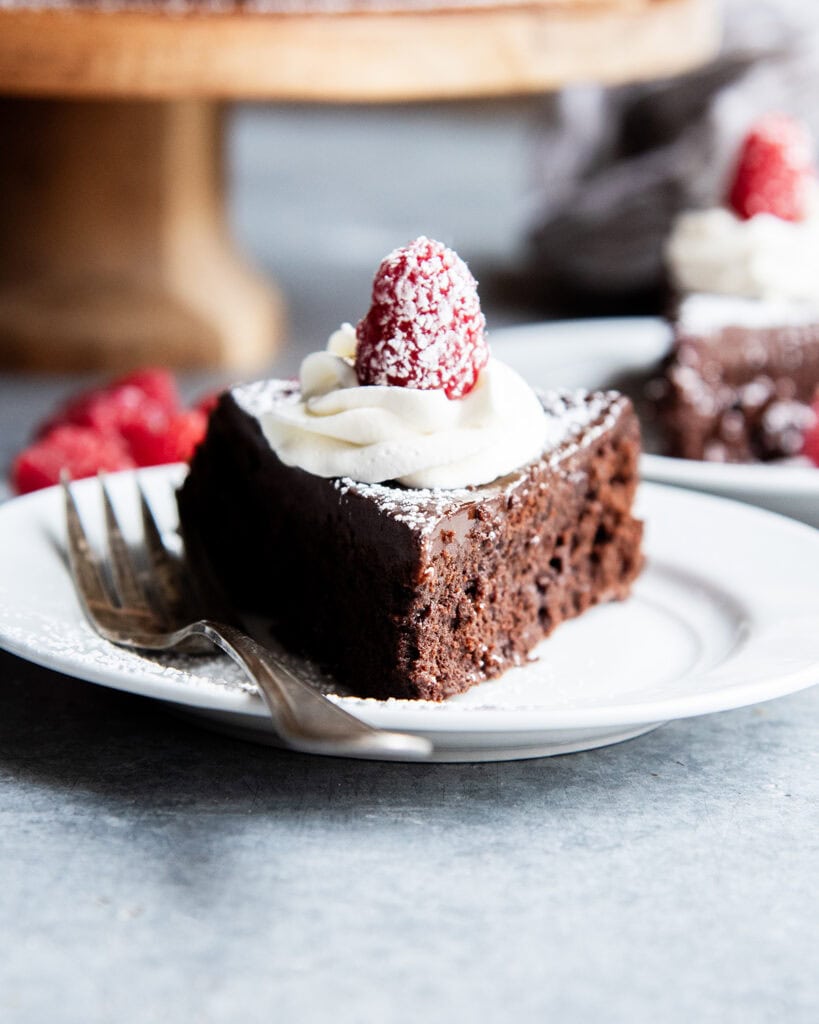 A slice of flourless chocolate cake topped with chocolate ganache, whipped cream, and a raspberry on a plate.