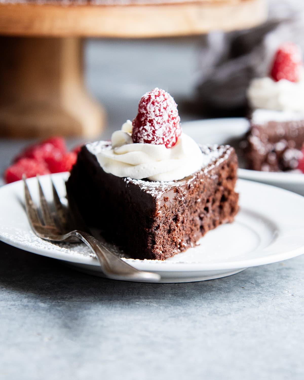 A slice of flourless chocolate cake topped with chocolate ganache, whipped cream, and a raspberry on a plate.