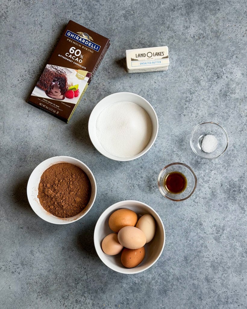 Chocolate bars, butter, sugar in a white bowl, eggs, cocoa powder, vanilla, and salt all the ingredients needed to make flourless chocolate cake. 