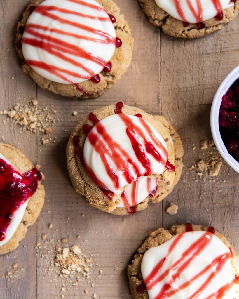 A wooden table topped with Raspberry cheesecake cookies with cream cheese frosting, and a drizzle of raspberry sauce on top.
