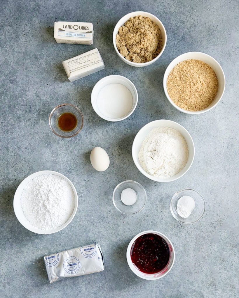 The ingredients needed to make Raspberry Cheesecake Cookies in small bowls including brown sugar, graham cracker crumbs, flour, raspberry jam, etc. 