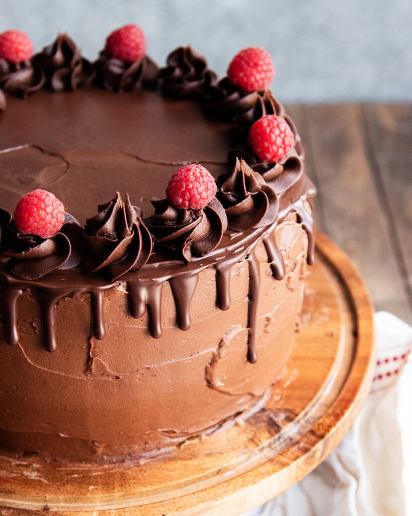A three layer chocolate raspberry cake on a wooden cake stand, topped with fresh raspberries.