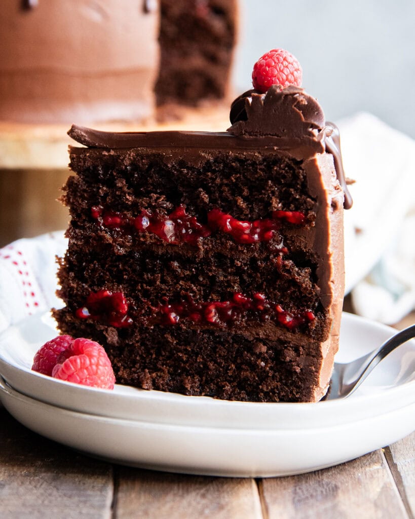 A slice of triple layer chocolate raspberry cake standing on a plate. It's topped with a dollop of chocolate buttercream, and a fresh raspberry.
