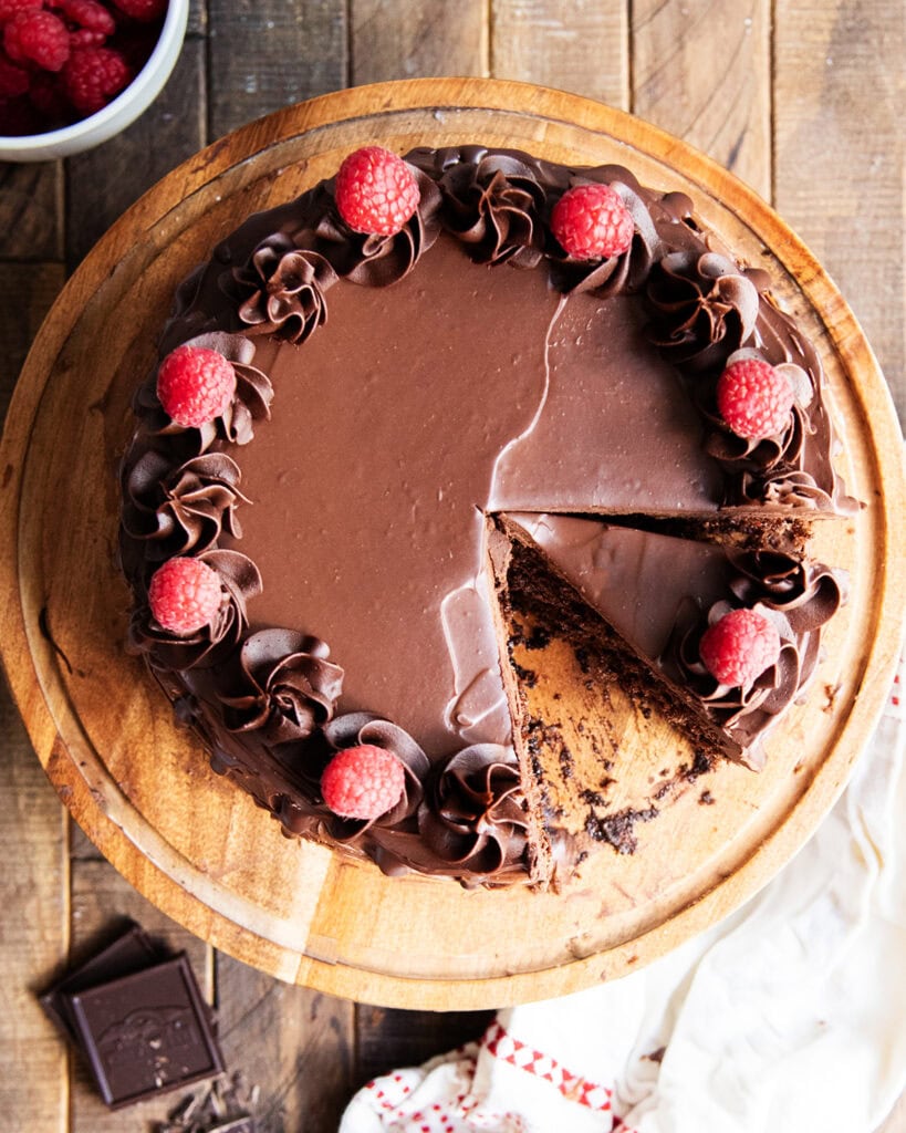 A round chocolate cake on a wooden cake stand topped with dollops of chocolate buttercream and fresh raspberries, one slice is cut out of the cake.
