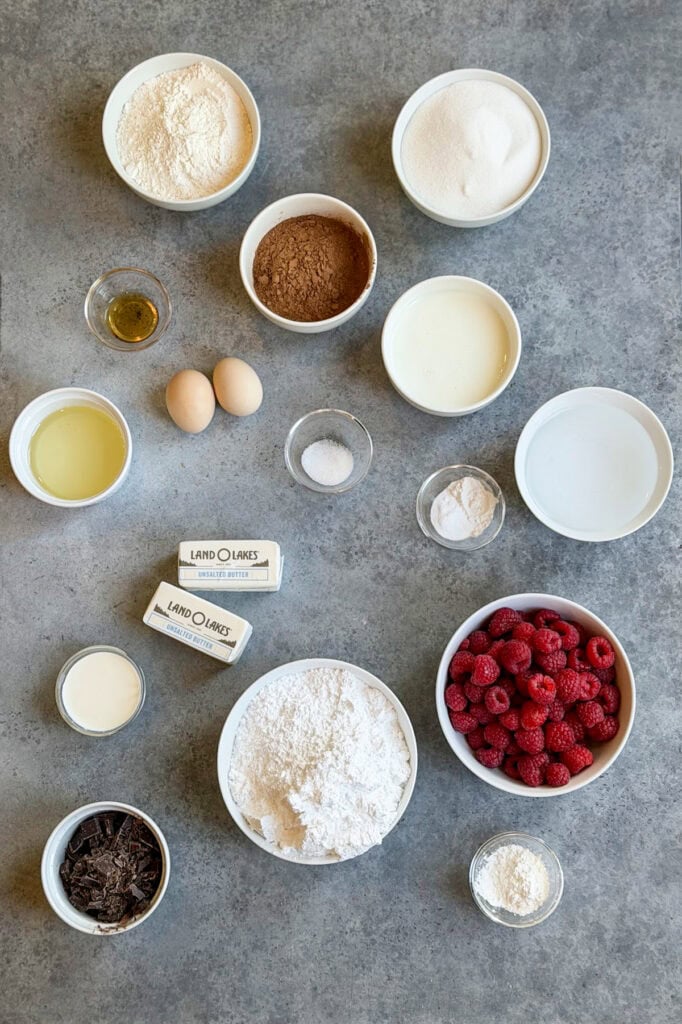 Small bowls of ingredients needed to make chocolate raspberry cake, there is fresh raspberries, cocoa powder, flour, powdered sugar, eggs, salt, butter, and more.