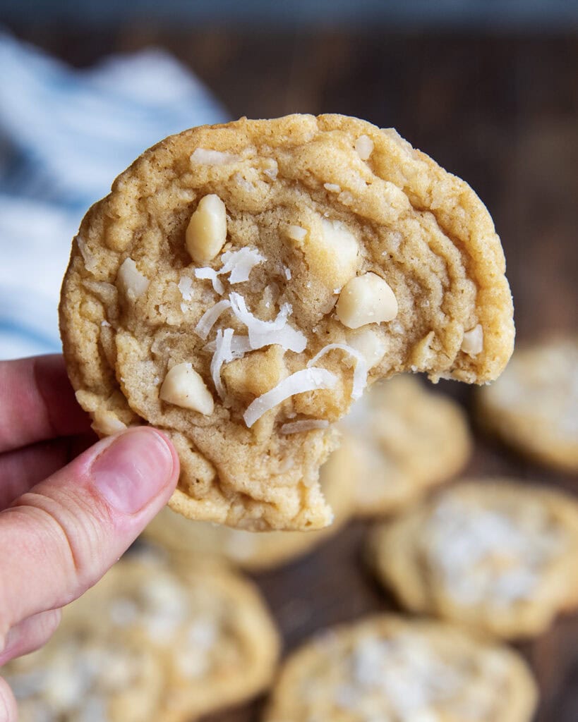 A hand holding a coconut macadamia nut cookie with a bite out of it.