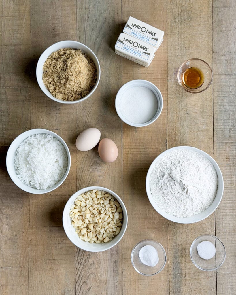 Small bowls of the ingredients needed to make coconut macadamia nut cookies, like brown sugar, butter, eggs, coconut, flour, and macadamia nuts. 