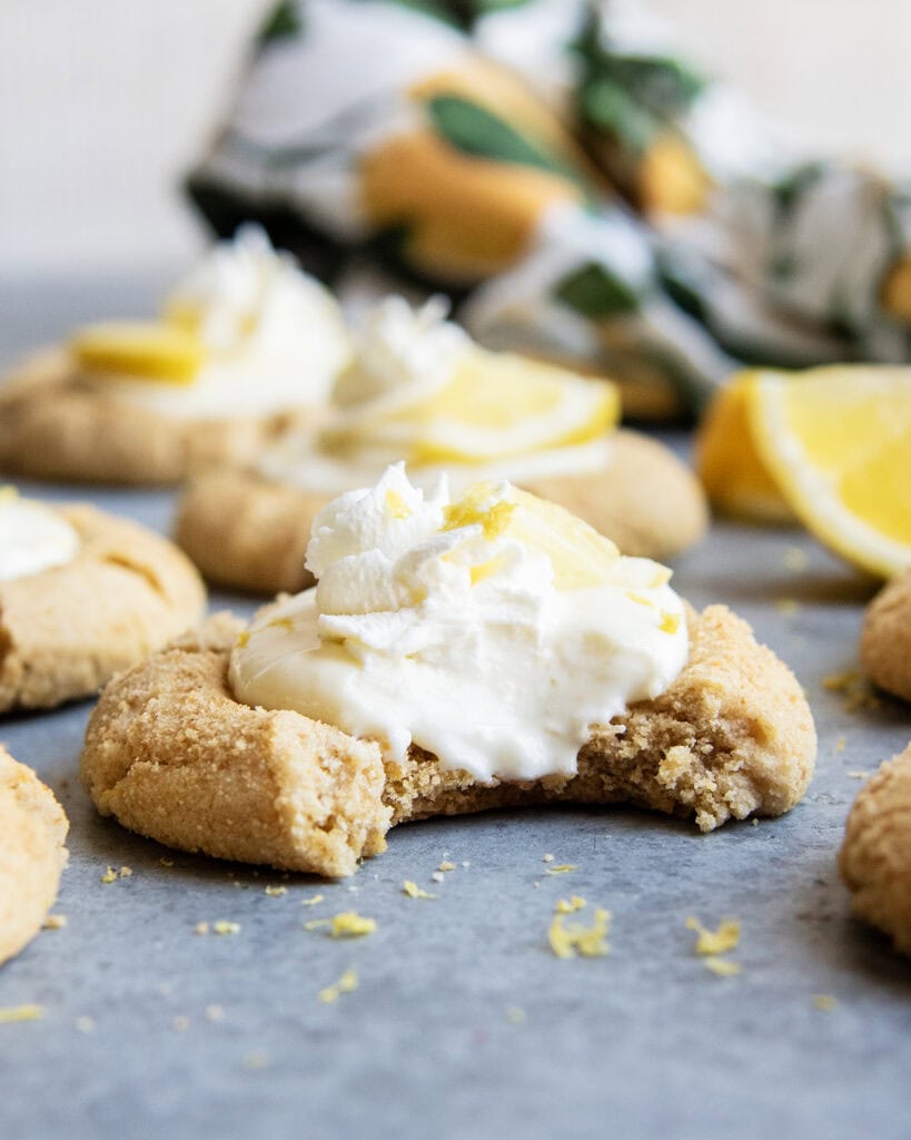 Lemon cream pie cookies with whipped cream and a lemon wedge on top on the counter, the front cookie has a bite out of it.