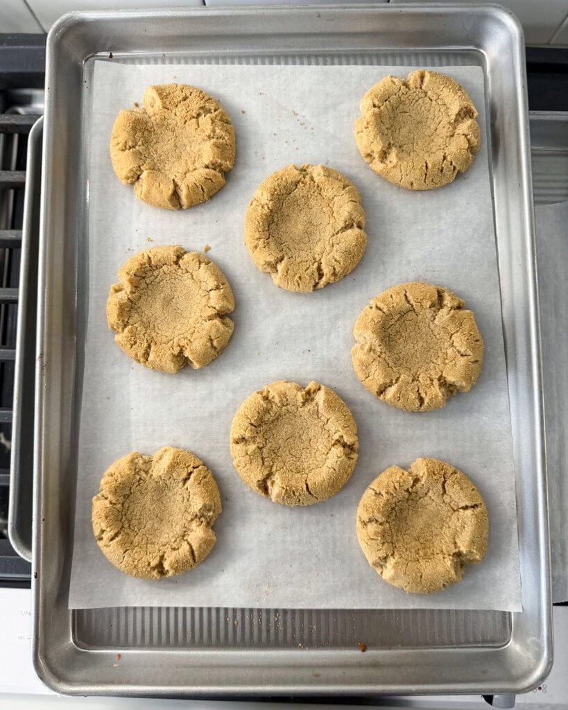 Crackled cookies with an indent pressed in the middle on a cookie sheet.