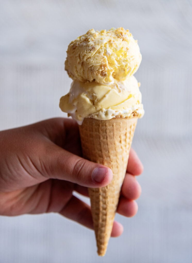 A hand holding an ice cream cone with two scoops of lemon cream pie ice cream on a sugar cone.
