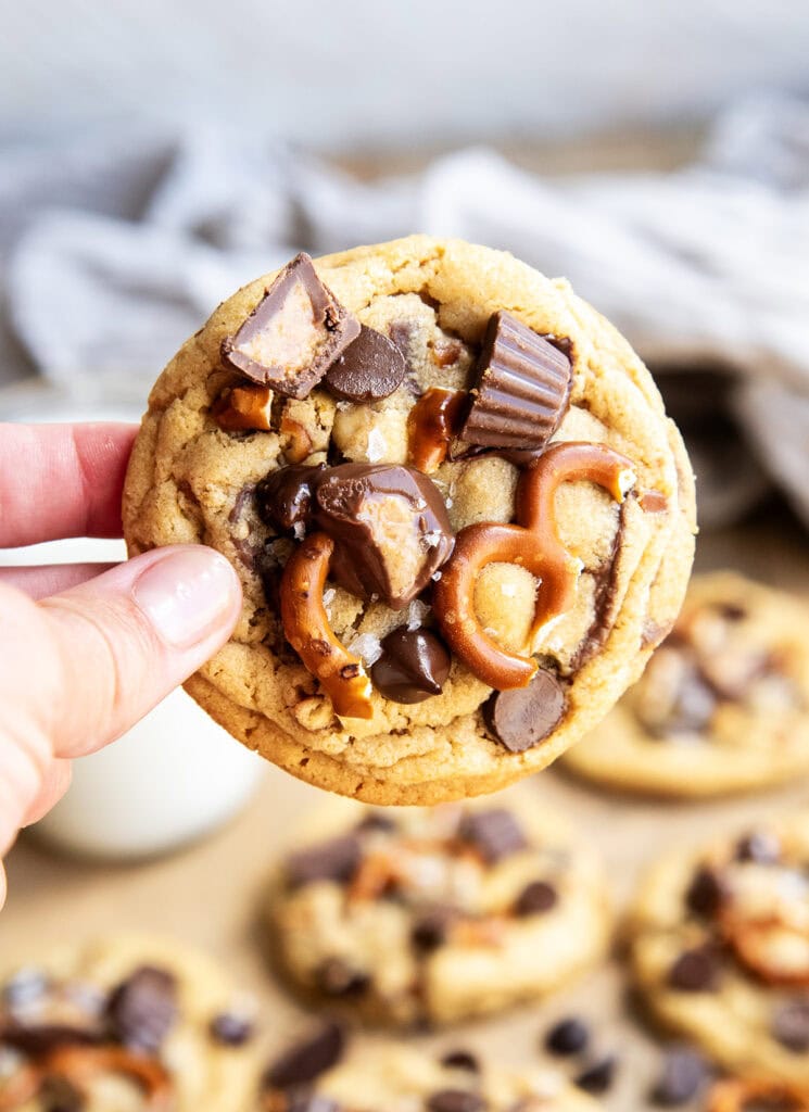 A hand holding a peanut butter pretzel cookie topped with mini peanut butter cups, and pretzels, and chocolate chips.