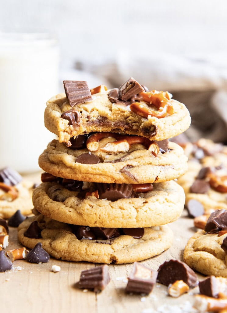 A stack of four peanut butter pretzel cookies topped with mini pretzels, and peanut butter cups, the top cookie has a bite out of it.