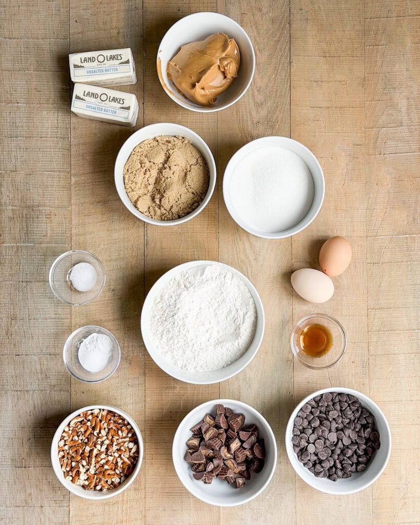 The ingredients needed to make peanut butter pretzel cookies, in small bowls. Like peanut butter, brown sugar, eggs, pretzels, chocolate chips, and flour.