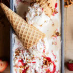 Two cheesecake ice cream cones on a pan of the ice cream, with strawberry swirls in the ice cream.
