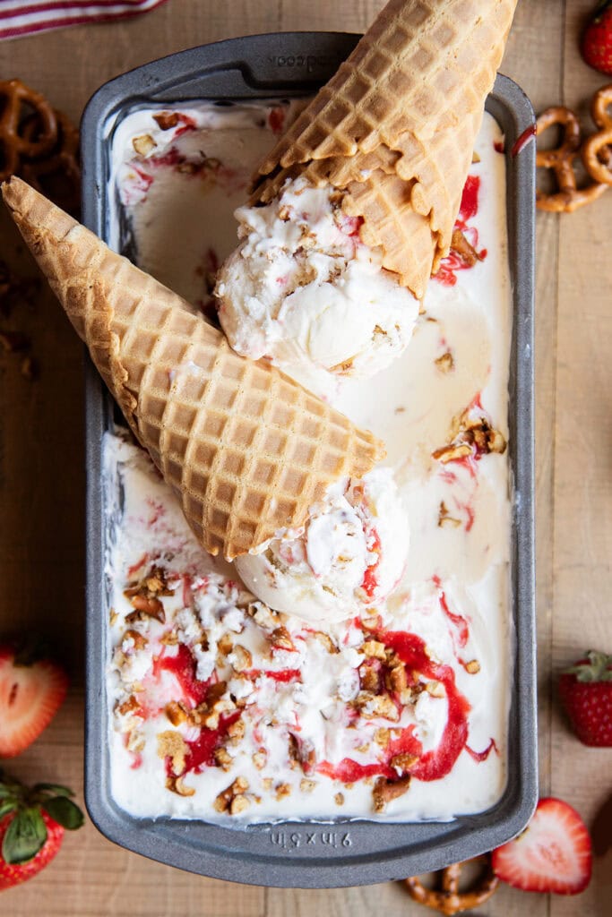 Two cheesecake ice cream cones on a pan of the ice cream, with strawberry swirls in the ice cream.
