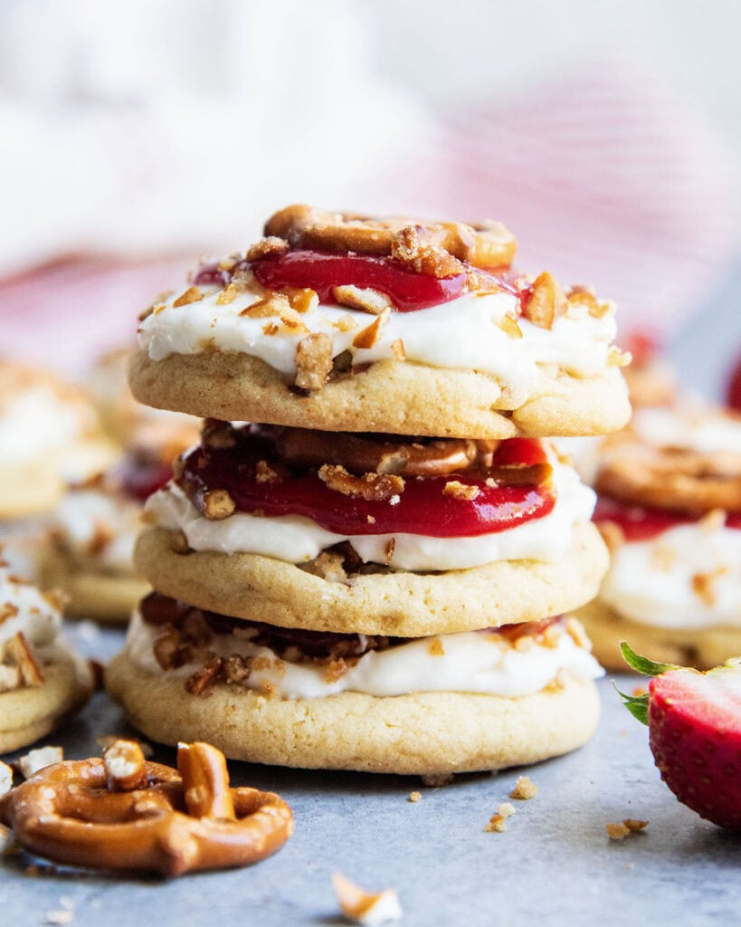 A stack of three strawberry pretzel salad cookies, each cookie is topped with whipped cream cheese frosting, strawberry sauce, and a pretzel.