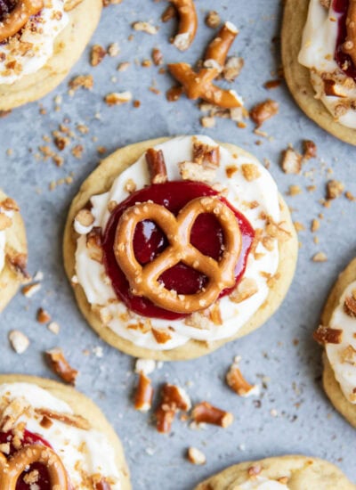 An above view of a cookie topped with cream cheese frosting, strawberry sauce and a mini pretzel.