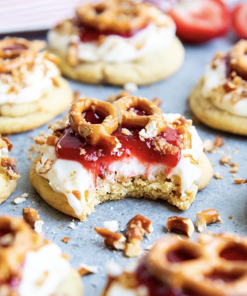 A strawberry pretzel salad inspired cookie with a cream cheese frosting, strawberry sauce, and mini pretzel. A bite is taken out of the cookie.