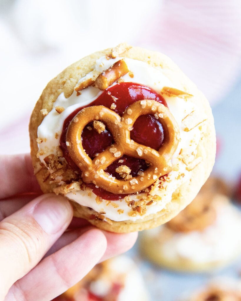 A hand holding a cookie topped with cream cheese frosting, strawberry sauce and a mini pretzel.