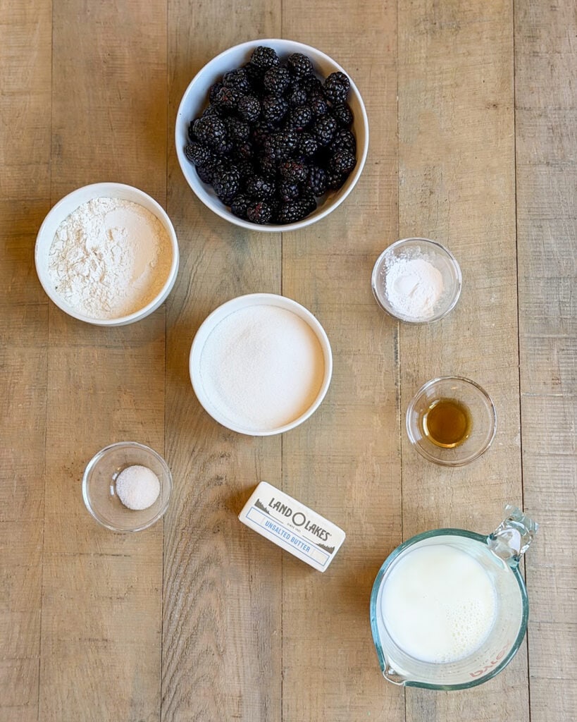 The ingredients needed to make blackberry cobbler in small bowls, you'll have blackberries, flour, butter, granulated sugar and vanilla.