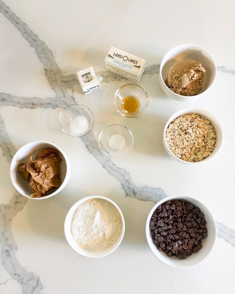 Small bowls of the ingredients needed to make cookie butter bars, like flour, oats, chocolate chips, brown sugar, and cookie butter.